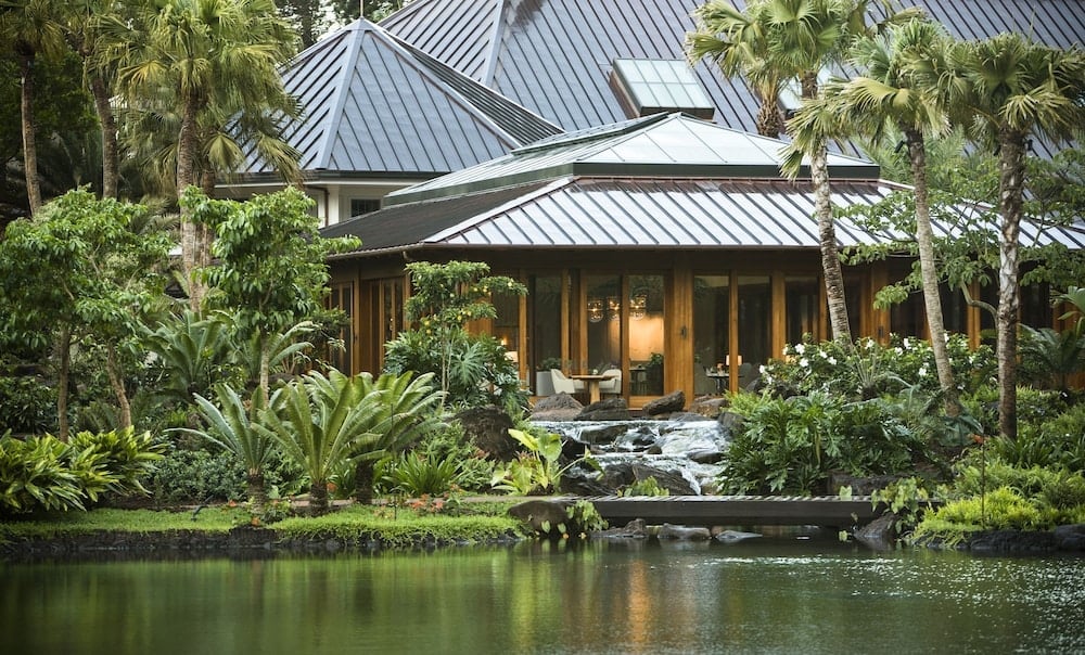 Volcanic stone pathways wind past koi ponds to private villa sanctuaries, where warm wood and metal rooflines disappear beneath swaying palms and tropical abundance.
