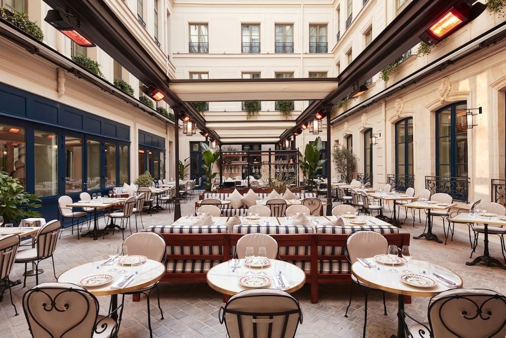 Striped banquettes and wrought-iron chairs create Parisian bistro charm in this elegant courtyard restaurant, where soft lighting and classical architecture frame intimate dining moments.