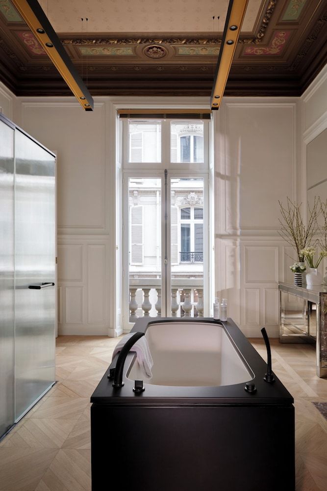 Ornate frescoed ceilings meet sleek modern design in this stunning suite bathroom, where a sculptural black tub takes center stage beneath soaring historic details.