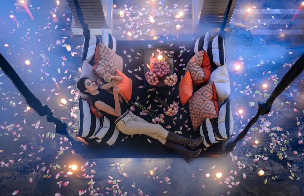 Rose petals cascade from glass ceiling panels above an intimate dining setup, creating a dreamlike underwater restaurant experience bathed in ethereal blue light.
