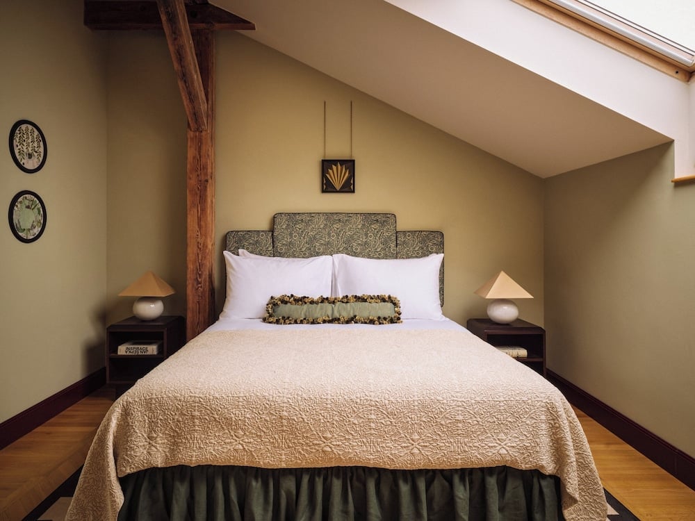 Exposed wooden beams frame this intimate attic sanctuary, where cream linens and sage green accents create a cocoon of rustic elegance beneath slanted skylights.