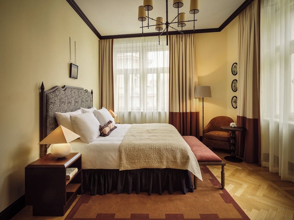 Warm amber light filters through gauze curtains, illuminating rich wood headboards and brass chandeliers that define Sir Prague's sophisticated Art Deco-inspired guest quarters.