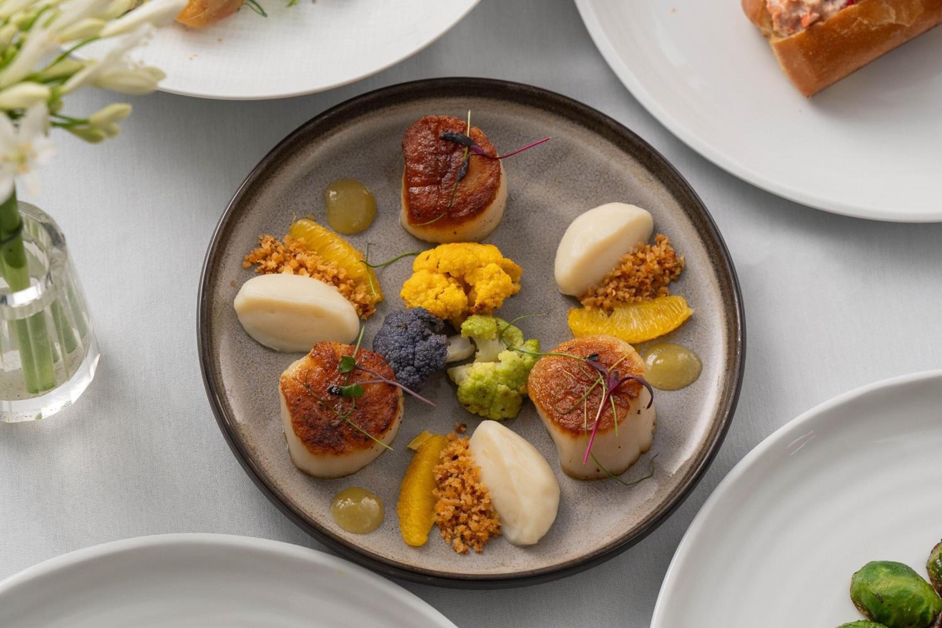 Seared scallops arrive artfully plated with vibrant cauliflower, citrus segments, and microgreens—a testament to the refined culinary artistry that defines Central Park South dining.