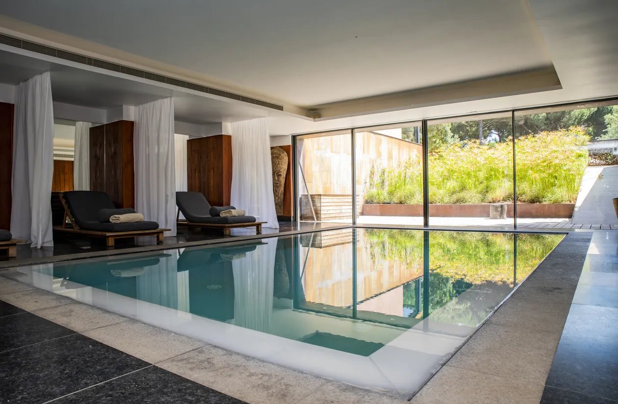 Sunlight streams through floor-to-ceiling windows into the serene spa pool, where draped daybeds and polished stone create an atmosphere of understated luxury.