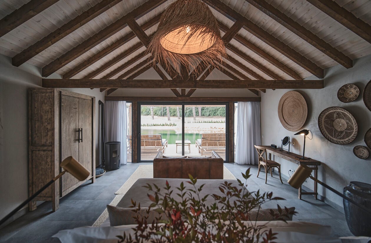 Exposed timber beams frame a serene suite where handwoven baskets adorn whitewashed walls, and a striking rattan pendant casts golden shadows across polished concrete floors.