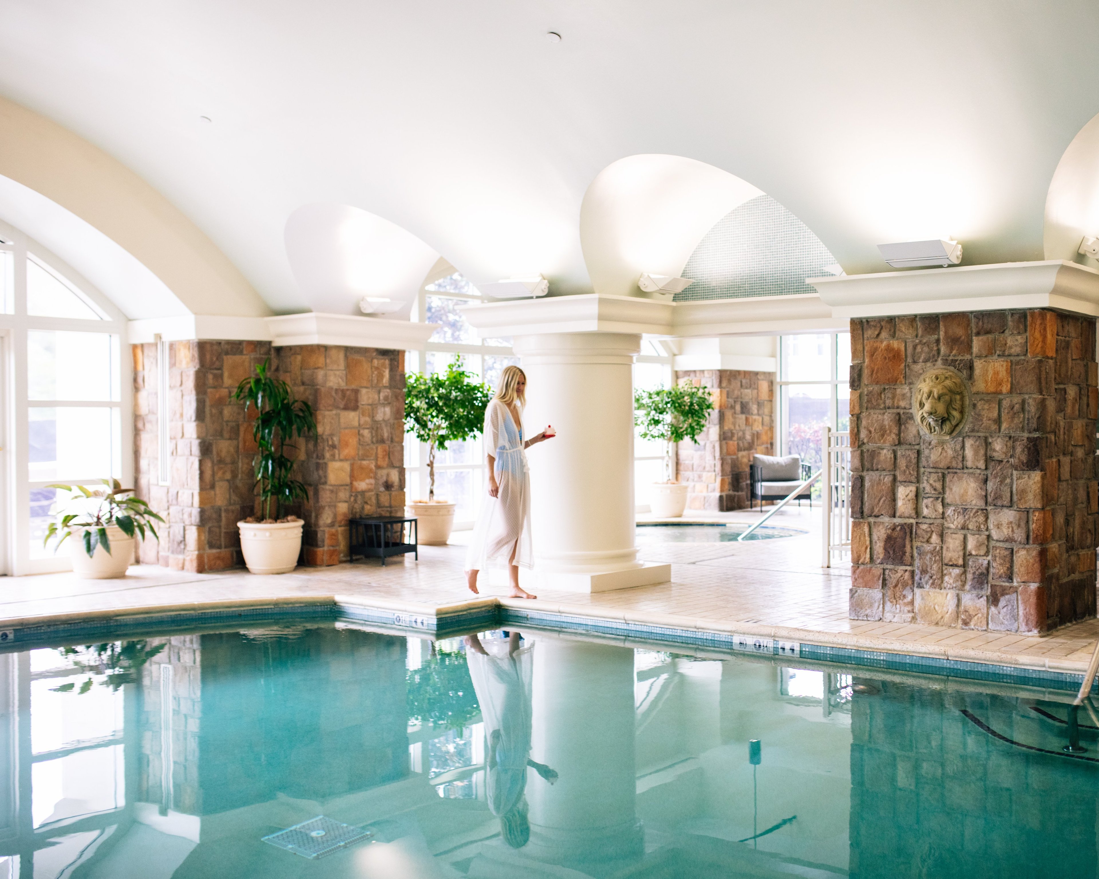 Natural light streams through arched windows onto the serene indoor pool, where stone columns and potted greenery create a tranquil sanctuary beneath soaring white ceilings.