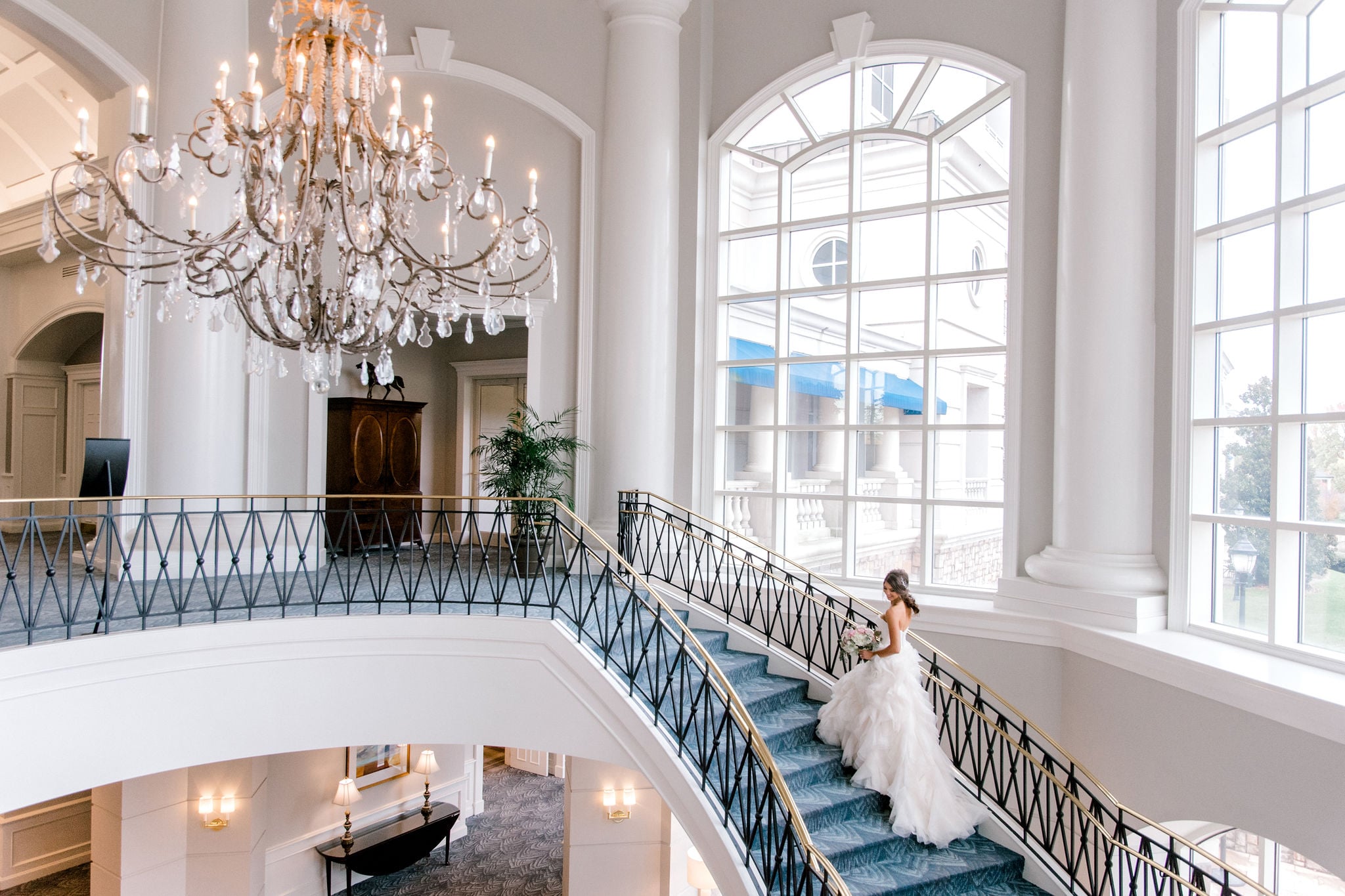 Crystal teardrops catch afternoon light as a bride descends the sweeping staircase, her gown trailing across blue carpet beneath soaring white columns.