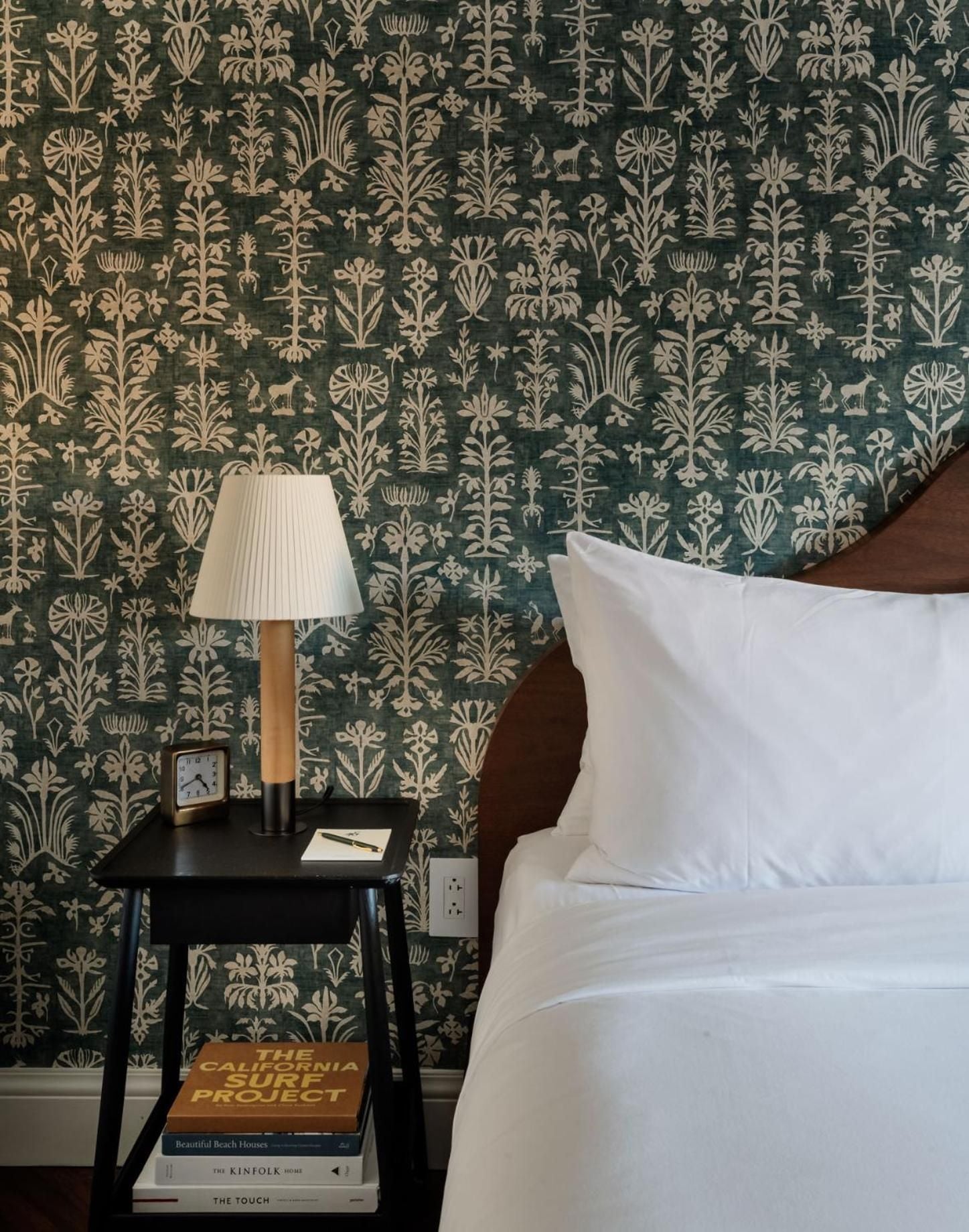Intricate botanical wallpaper in deep forest green creates an intimate backdrop for crisp white linens and mid-century bedside styling at this thoughtfully designed Charleston retreat.