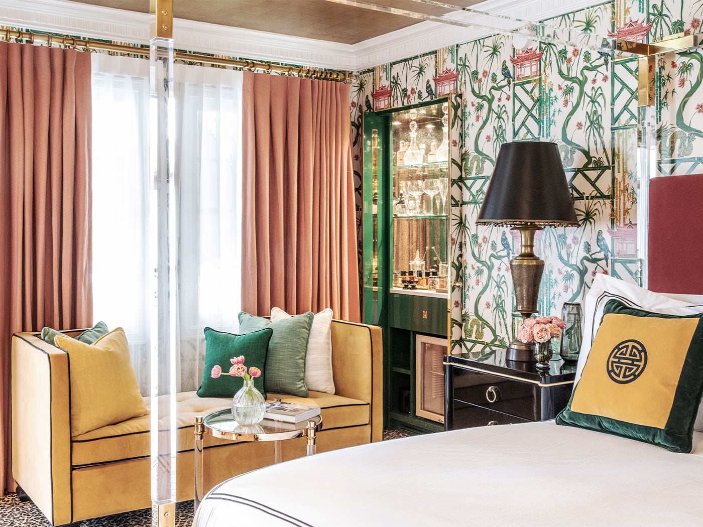 Emerald-green chinoiserie wallpaper and golden velvet seating create an intimate corner where peach curtains filter Hollywood light through oversized windows.