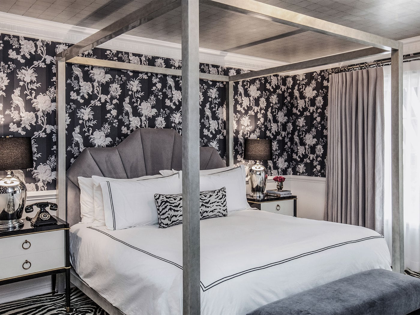 Dramatic black floral wallpaper envelops this canopy bed suite, where crisp white linens and zebra-print pillows create striking contrast against the moody botanical backdrop.