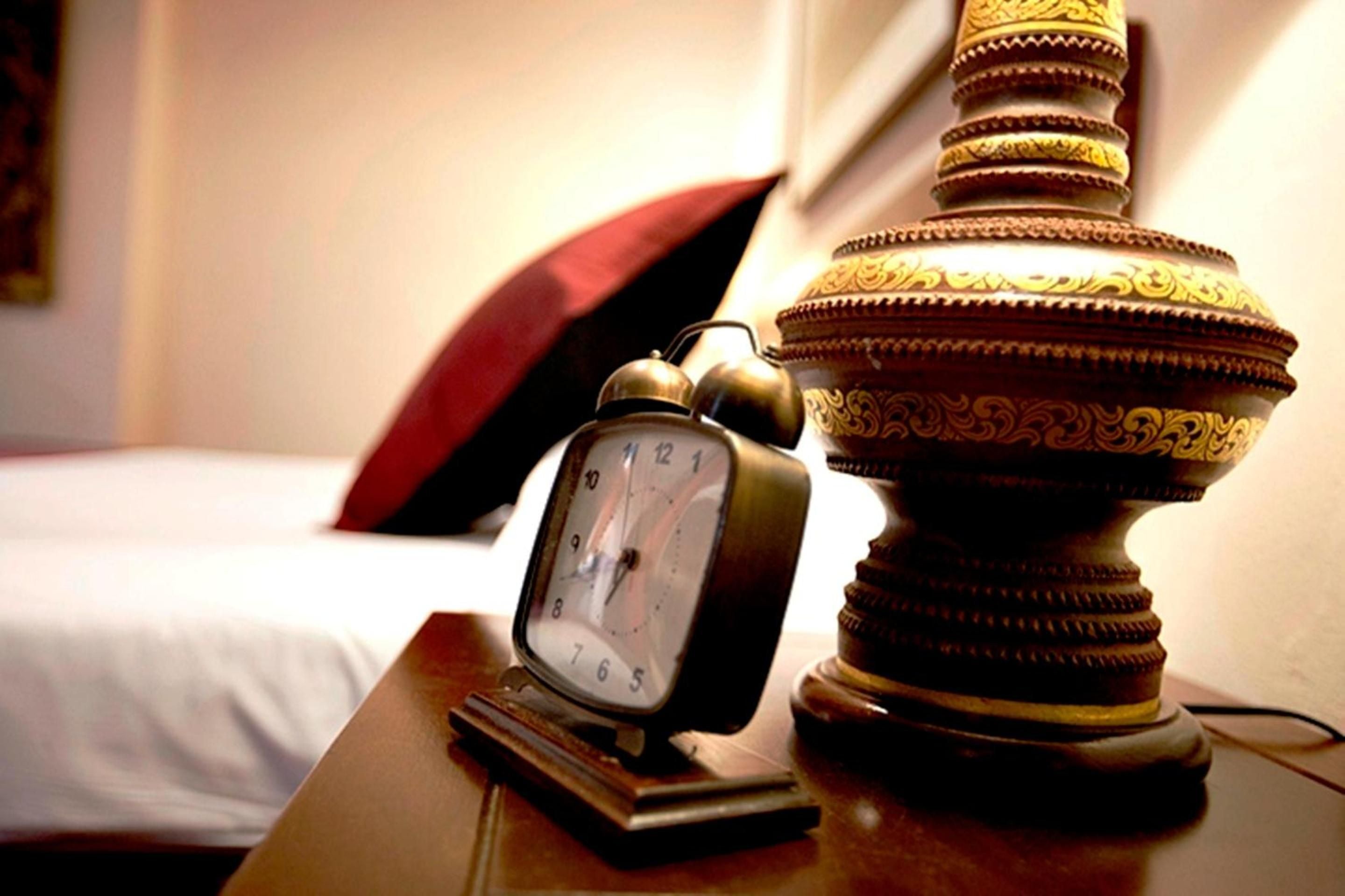 Ornate Thai artistry meets modern convenience on polished bedside tables, where vintage alarm clocks tick beside gilded ceremonial vessels in this culturally immersive suite.
