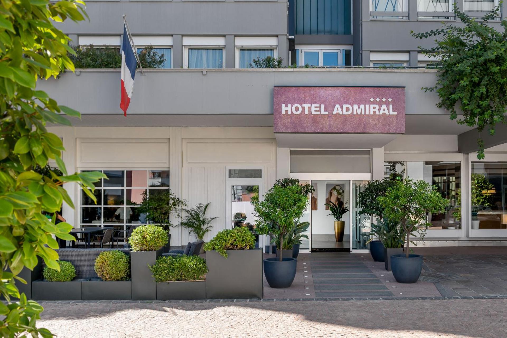 Crisp white façade and manicured boxwood planters create an inviting street presence, while the French tricolor signals sophisticated European hospitality at this polished four-star property.