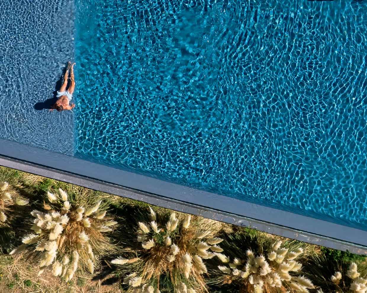 Crystal-blue waters shimmer beneath desert sun as a guest floats serenely, surrounded by native pampas grass swaying in Atacama's gentle breeze.