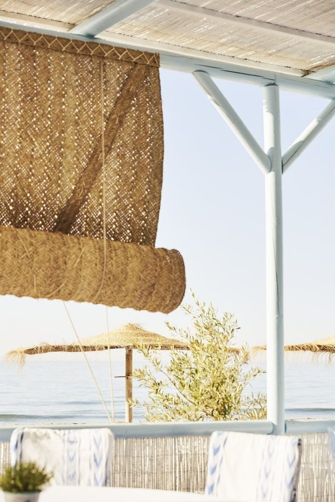 Woven bamboo screens flutter in Mediterranean breezes, framing sun-dappled terraces where natural textures create intimate pockets of shade overlooking the sparkling coastline.