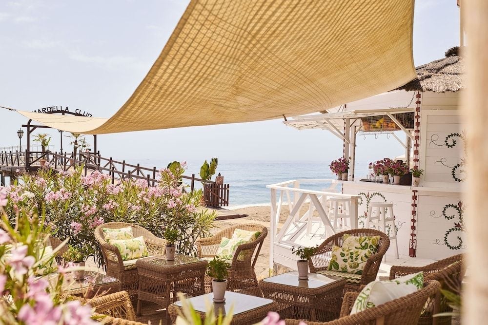 Wicker chairs and botanical cushions create an intimate terrace dining experience, where golden shade cloth filters Mediterranean light above blooming oleander and endless azure horizons.