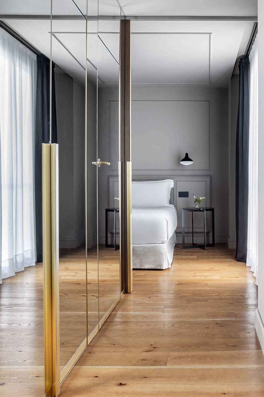 Glass partitions with brass frames create sophisticated transparency between living and sleeping spaces, while warm wood floors soften the contemporary gray palette.