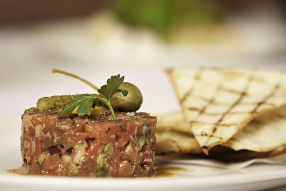Artfully composed tuna tartare arrives garnished with crisp capers and fresh herbs, accompanied by warm pita triangles that practically beg to be loaded with the silky raw fish.