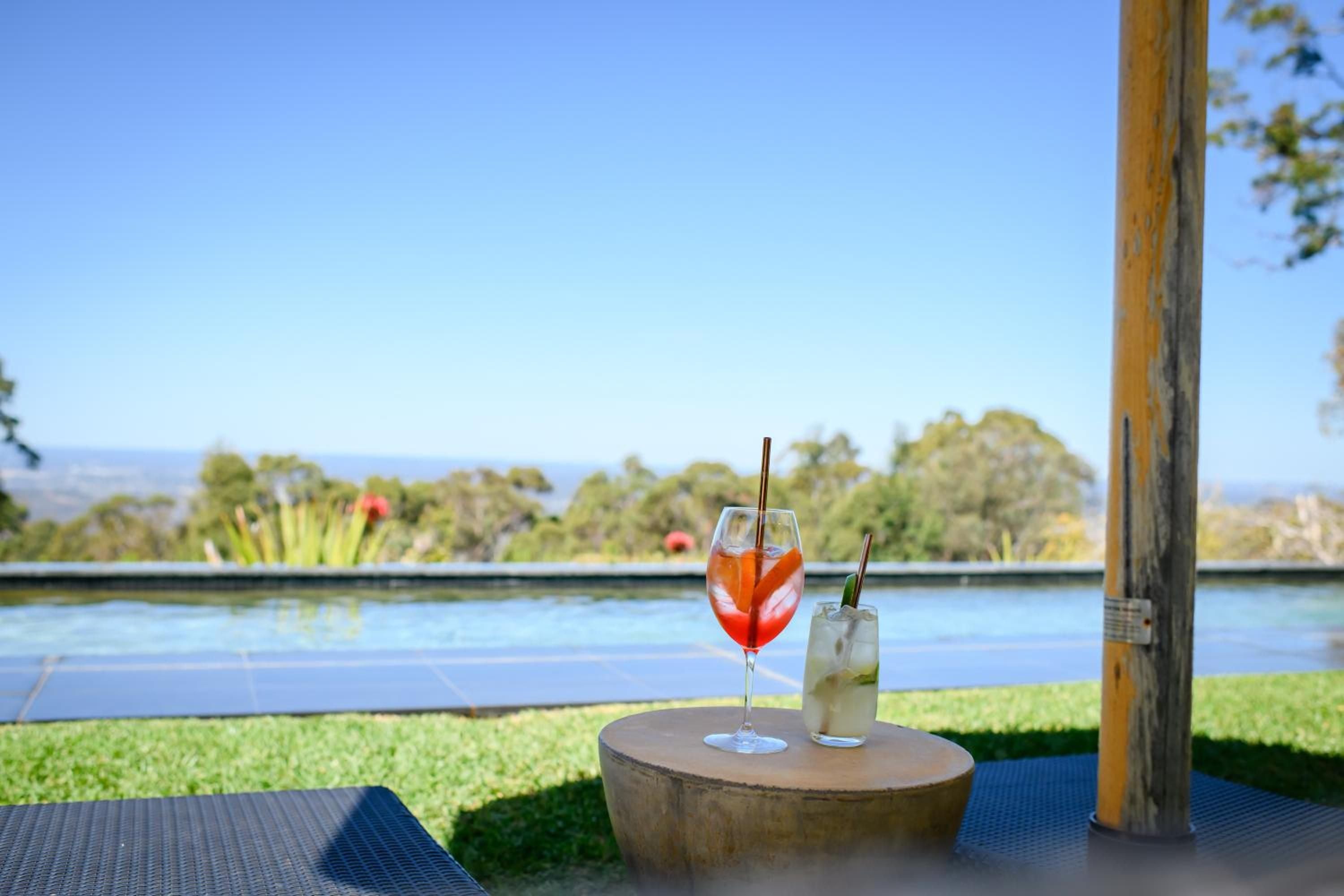 Handcrafted cocktails await on a wooden perch beside the infinity pool, where rolling hills dissolve into endless sky and afternoon light dances across still water.