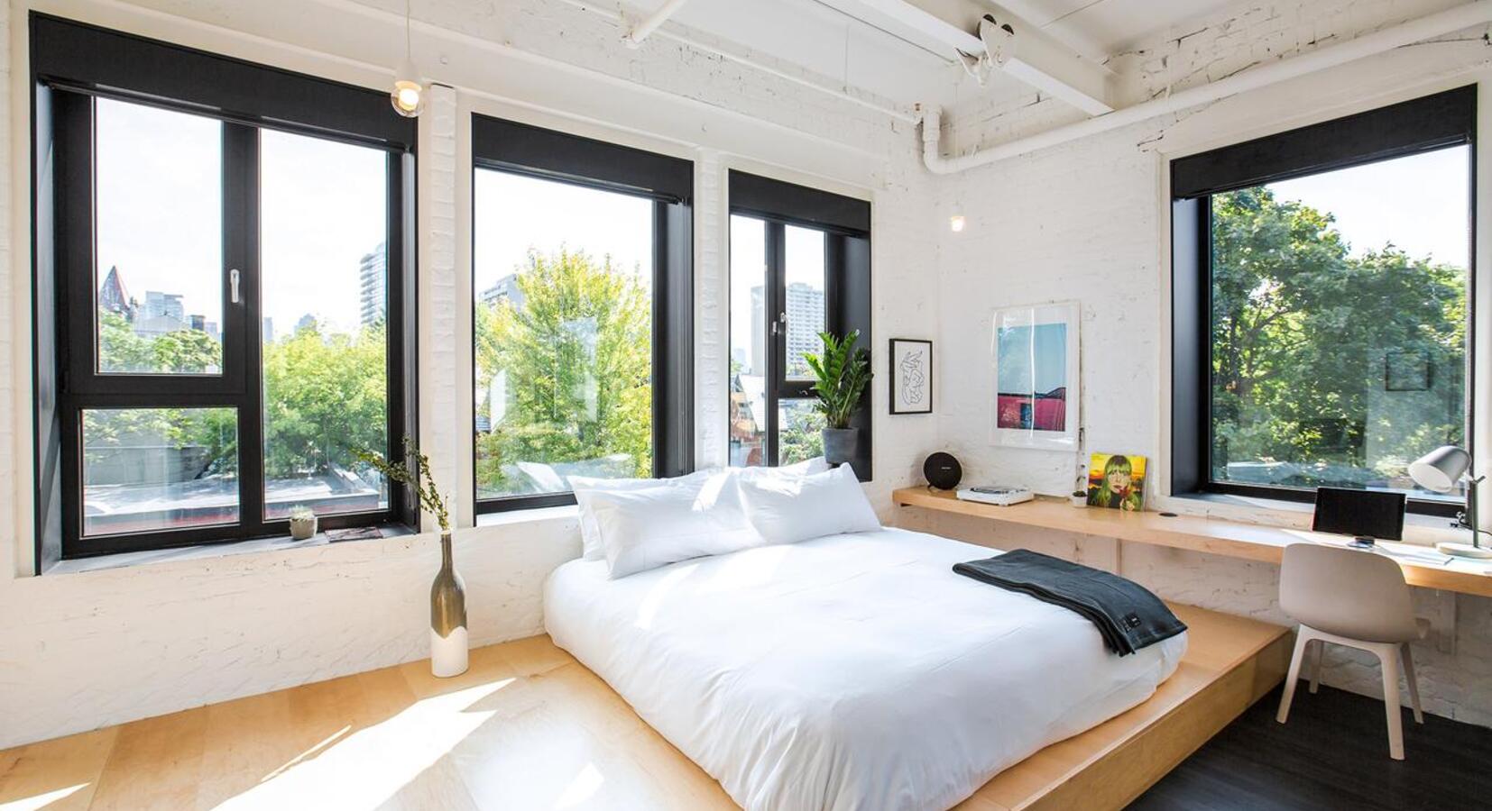 Floor-to-ceiling windows frame city skylines from this minimalist sanctuary, where exposed white beams meet warm wood platforms and crisp linens catch afternoon light.