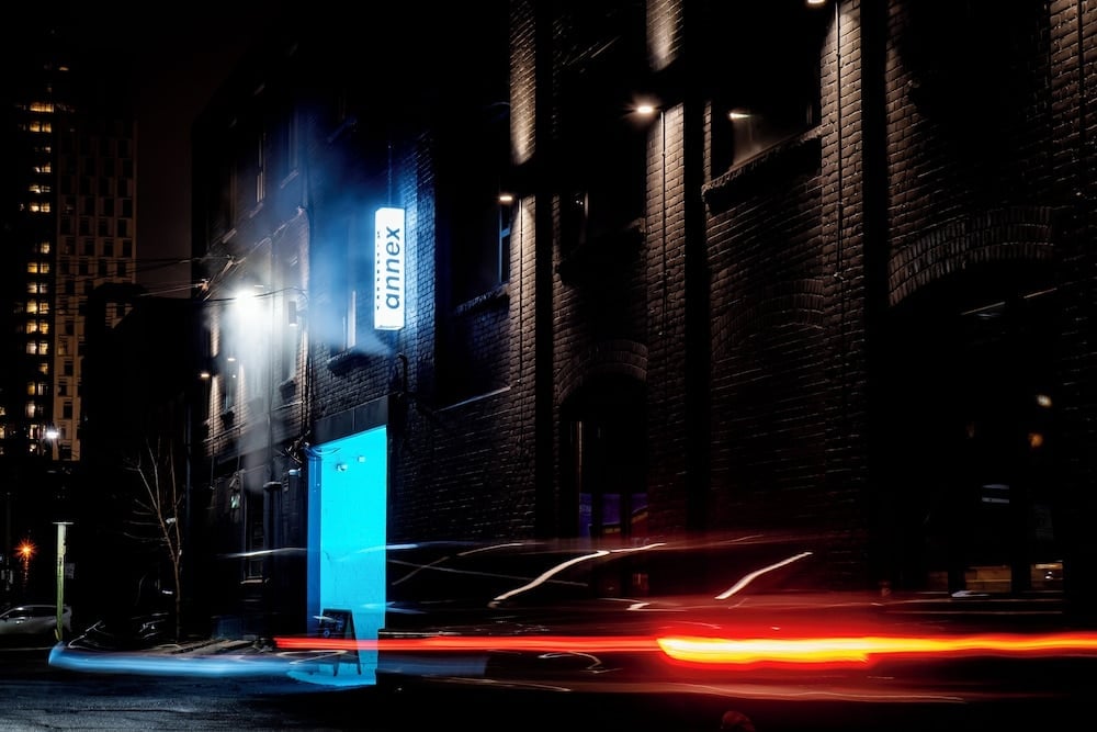 Illuminated signage cuts through the urban darkness, creating a beacon of contemporary hospitality against weathered brick walls as city lights streak past.
