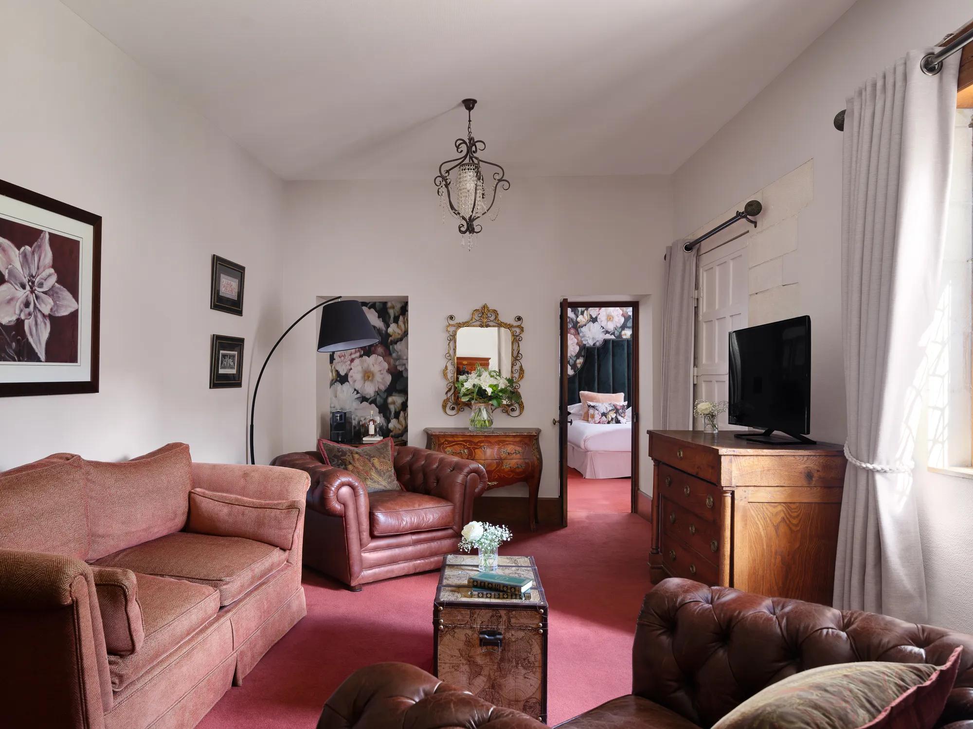 Antique furnishings and burgundy leather create an intimate salon where ornate mirrors catch afternoon light filtering through crisp white curtains in this refined suite.