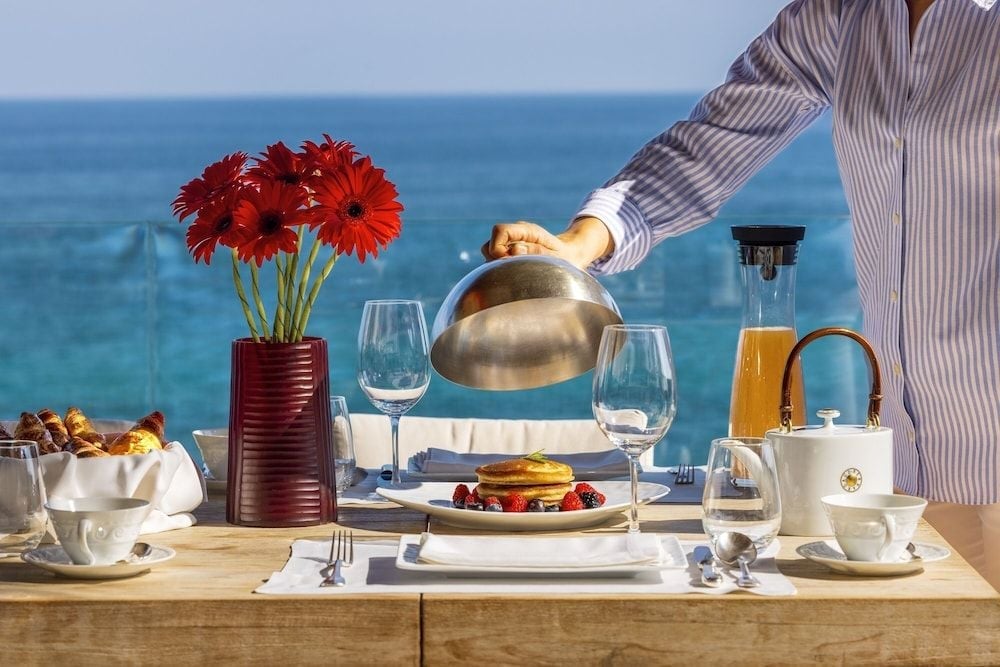 Mediterranean breakfast unfolds at your private table as azure waters stretch endlessly beyond, while fresh orange juice catches the morning light in this idyllic seaside setting.