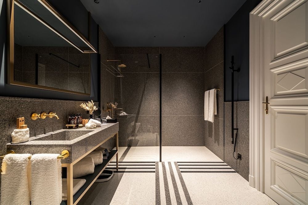 Dark terrazzo walls and brass fixtures create an intimate sanctuary where geometric floor patterns guide guests from vanity to rainfall shower in sophisticated harmony.