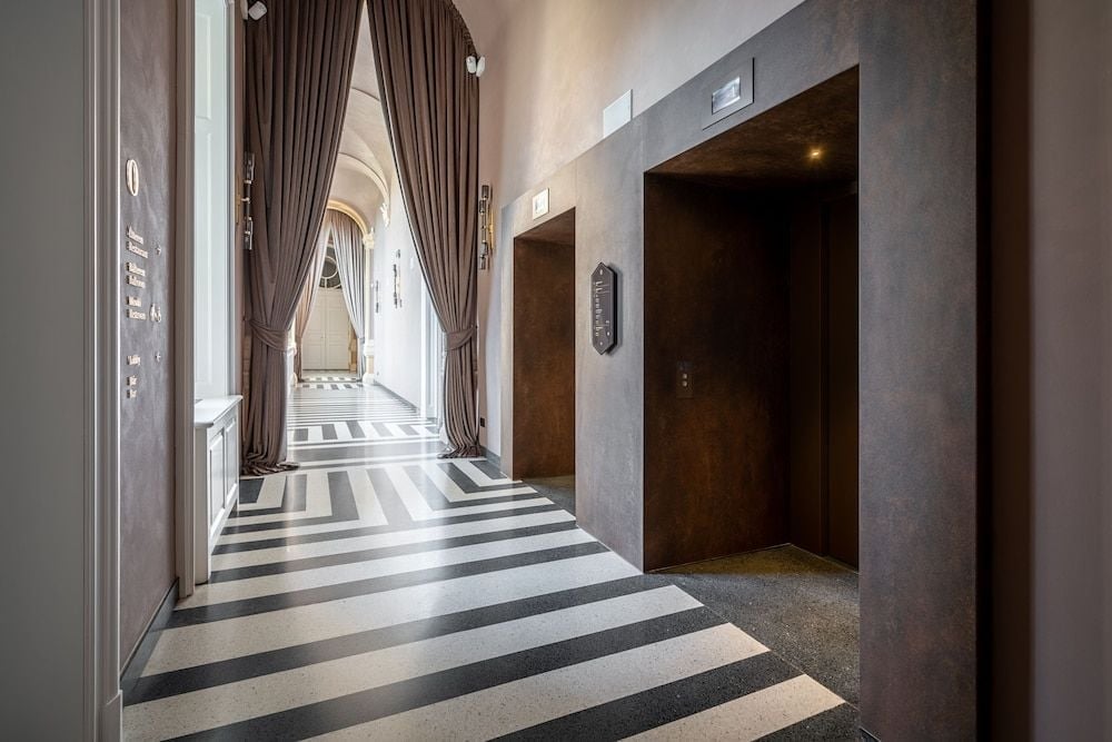 Dramatic black and white striped marble floors create a striking geometric pathway through elegant corridors, where rich brown curtains frame arched doorways and golden elevator doors await.