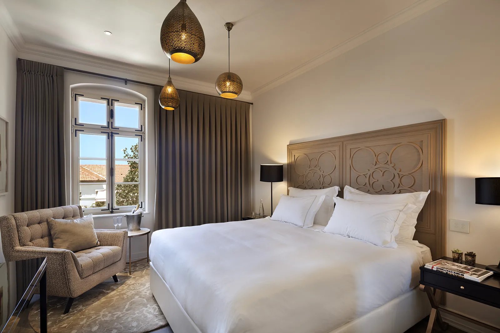 Moroccan-inspired pendant lights cast warm shadows over crisp white linens, while the carved wooden headboard adds Old World charm to this sophisticated guest room.