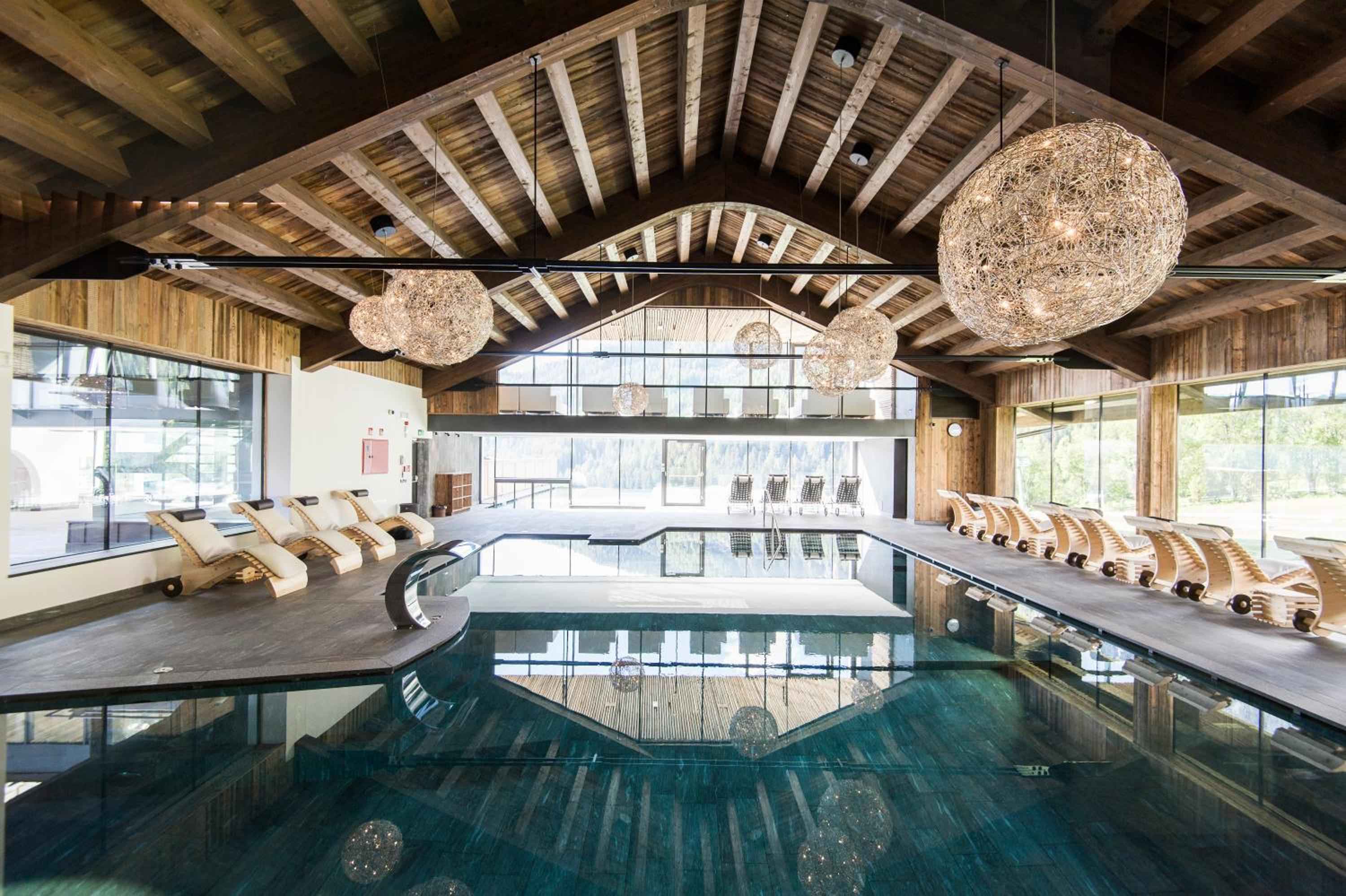 Suspended rattan orbs cast dappled shadows across the curved indoor pool, where exposed timber beams frame floor-to-ceiling windows overlooking alpine vistas.