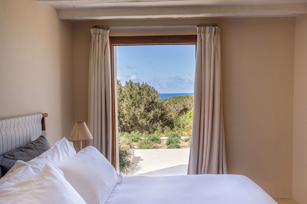 Mediterranean light streams through linen curtains, framing wild olive groves and azure coastline beyond crisp white linens and weathered wooden beams.