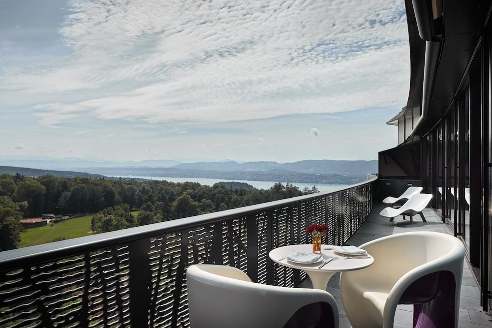 Curved white chairs frame intimate moments on a private terrace, where Lake Zurich stretches toward distant Alps beneath drifting clouds and golden afternoon light.