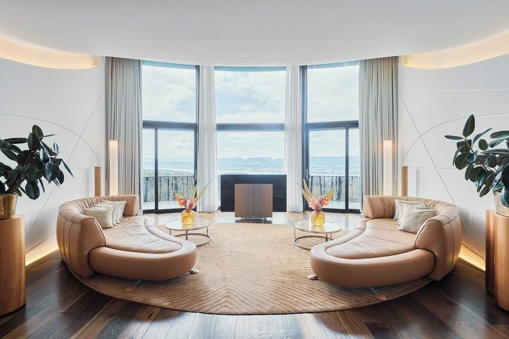 Curved cream leather sofas embrace floor-to-ceiling windows in this striking suite, where warm wood floors meet panoramic Alpine vistas through dramatic curved glass.