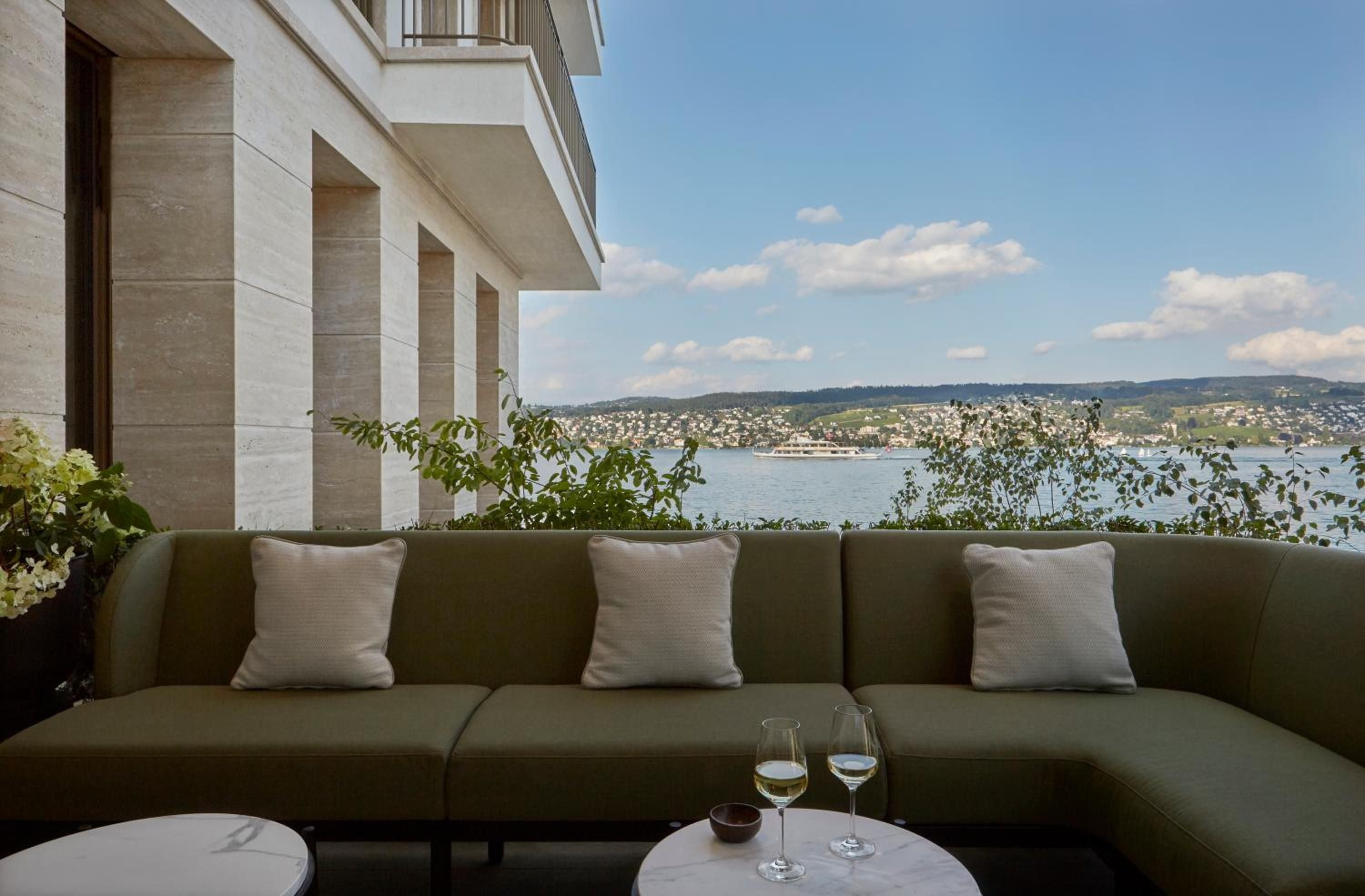 Olive-green cushions frame Lake Zürich's shimmering waters from this intimate terrace, where white wine catches afternoon light against rolling Swiss hillsides.