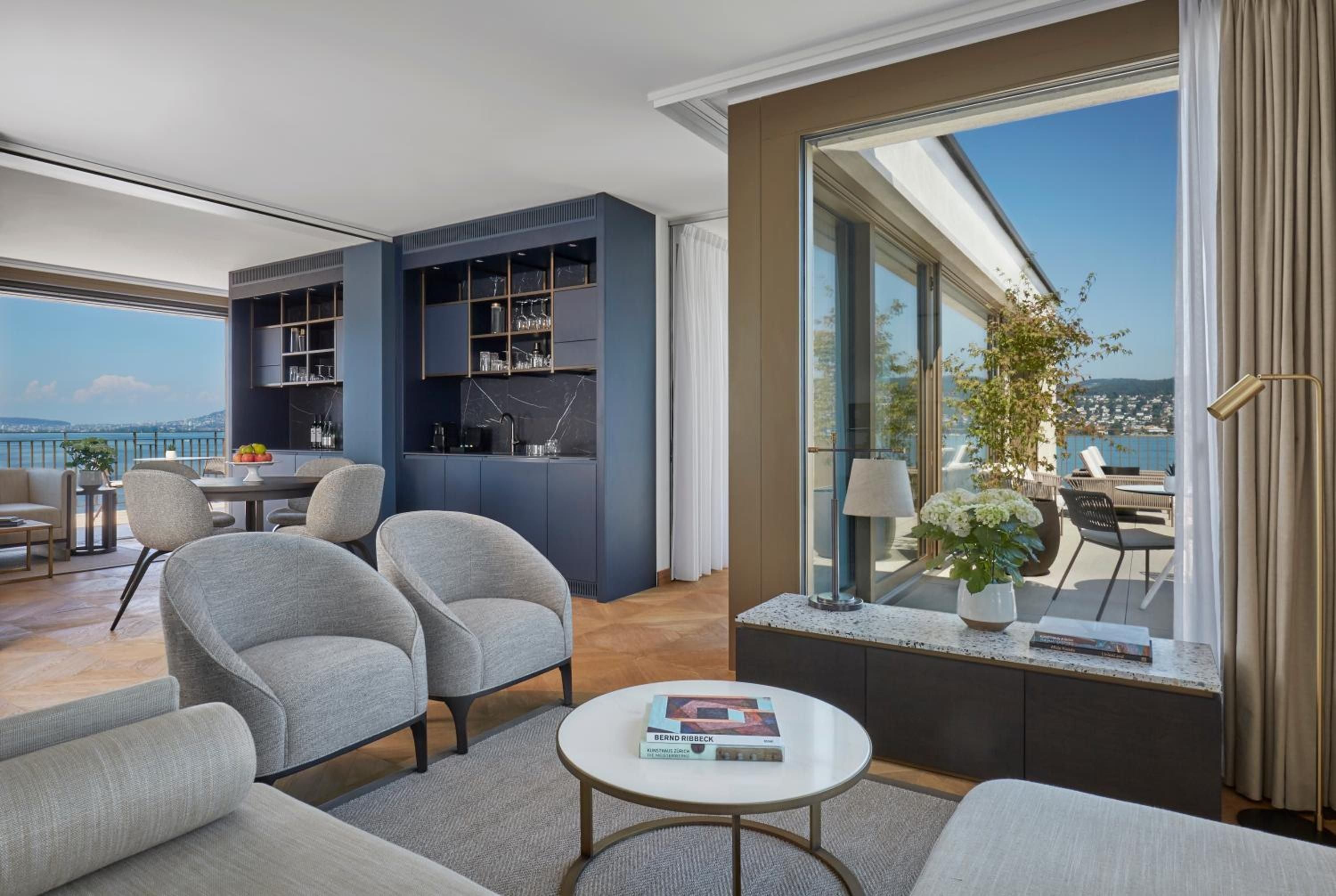Floor-to-ceiling windows frame Lake Zürich's azure waters from this sophisticated suite, where navy built-ins and soft gray seating create an effortlessly elegant retreat.