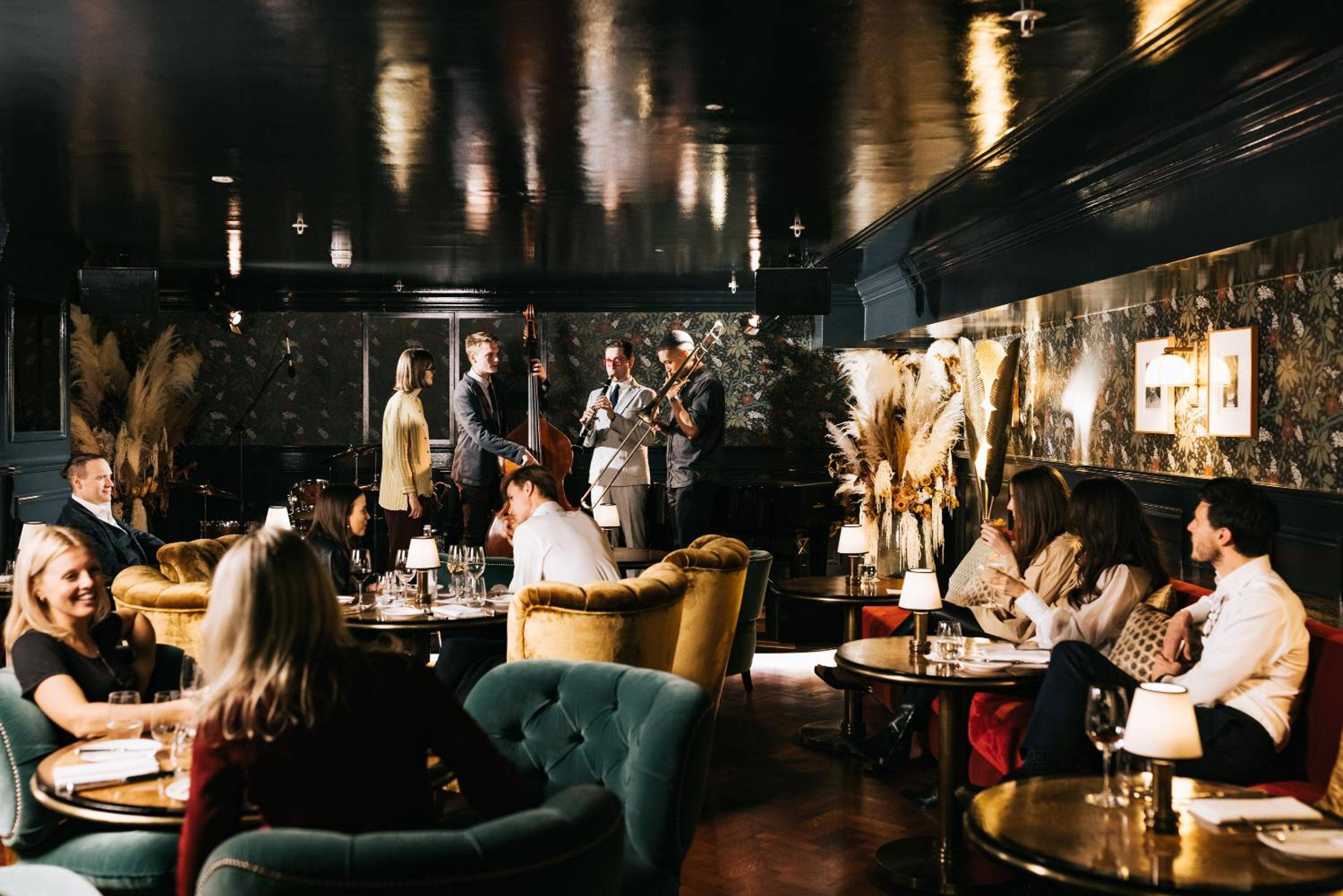 Jazz musicians serenade guests in Ned's Club's sumptuous lounge, where emerald velvet chairs and dramatic black walls create an intimate speakeasy atmosphere beneath gleaming pendant lights.