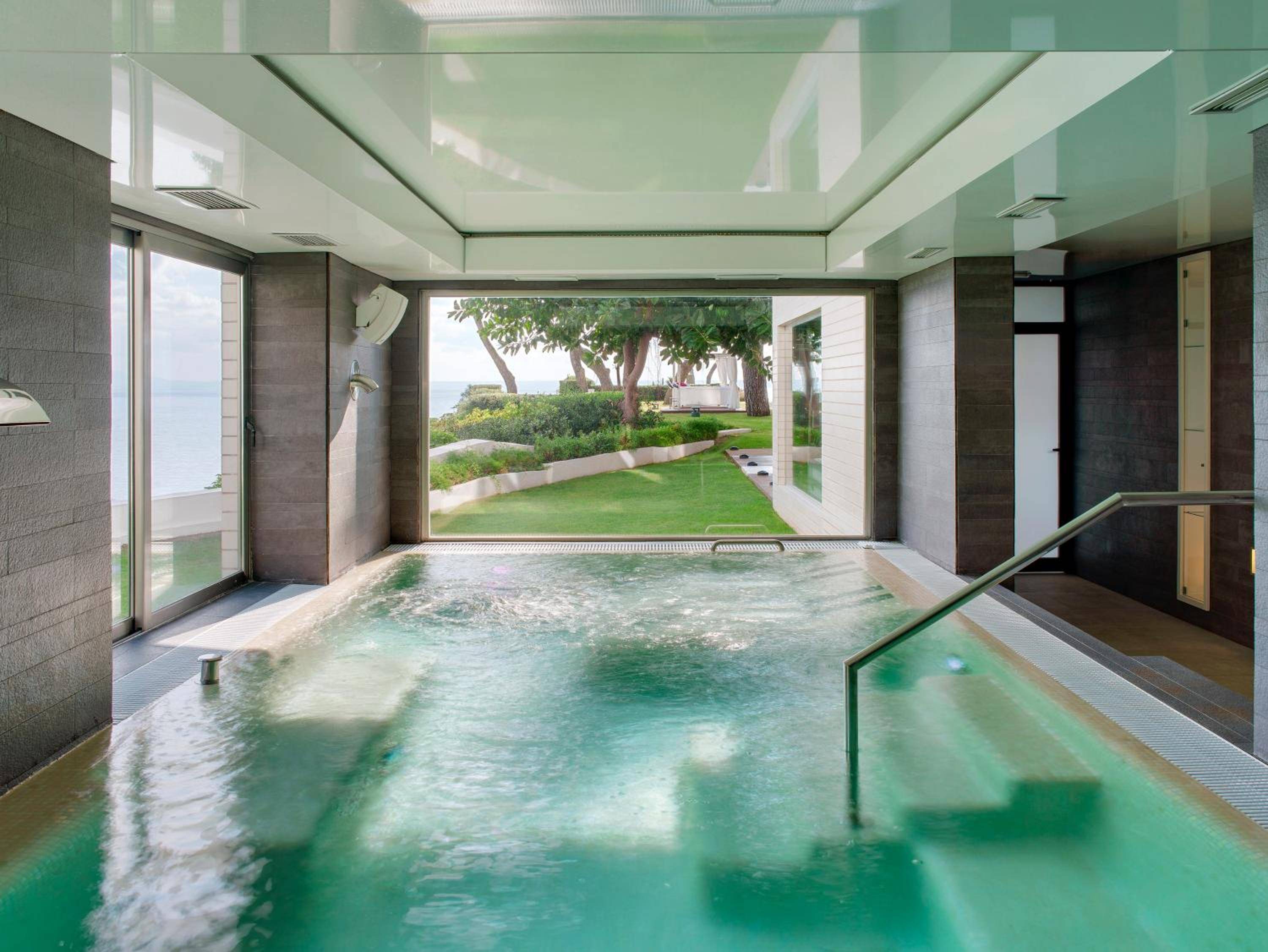 Steam rises from the jade-tinted hydrotherapy pool as floor-to-ceiling windows frame manicured gardens, creating a serene sanctuary where wellness meets Mediterranean elegance.