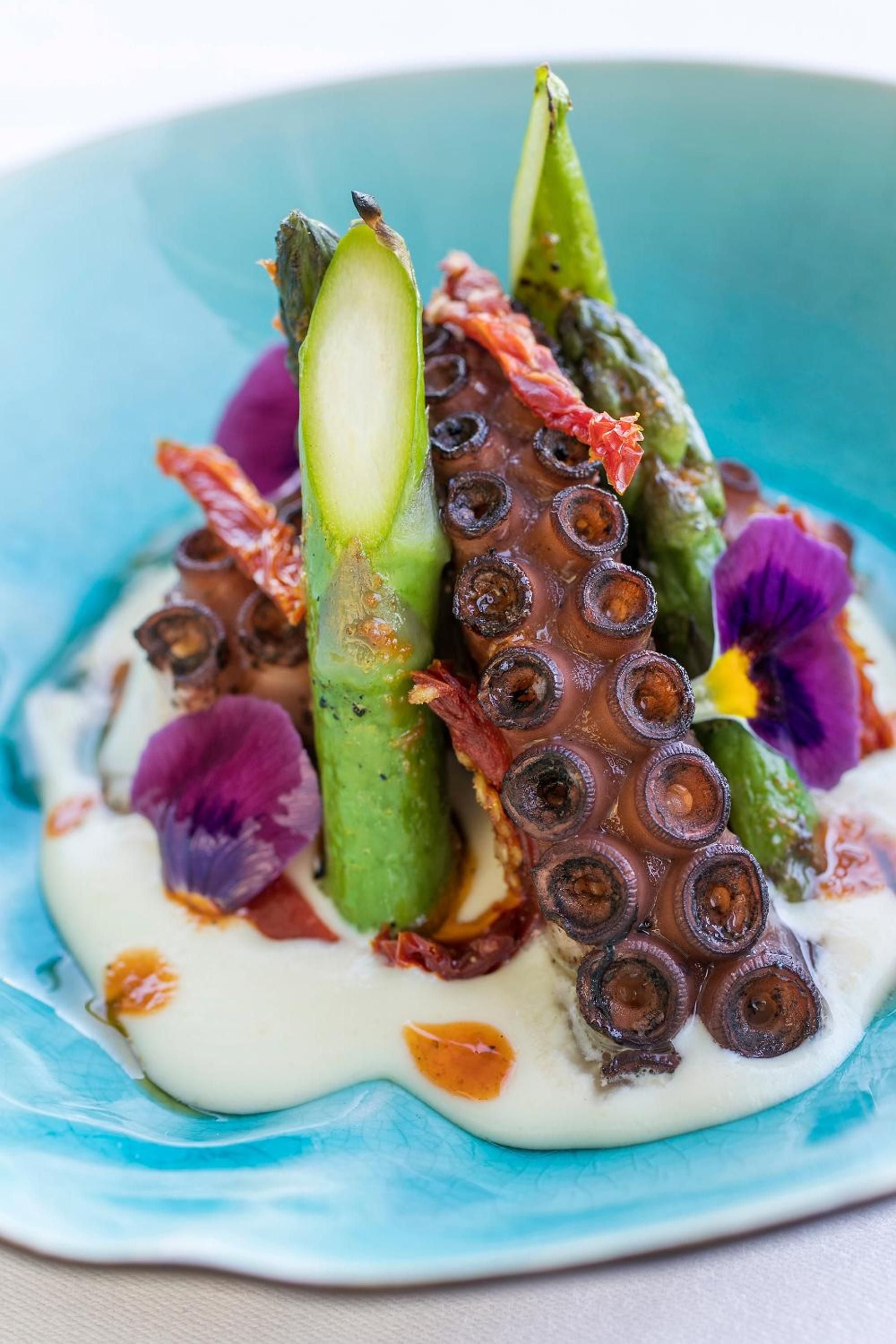 Artfully plated octopus tentacles crowned with vibrant asparagus spears and purple pansies showcase the culinary artistry that defines fine dining experiences at this sophisticated coastal retreat.