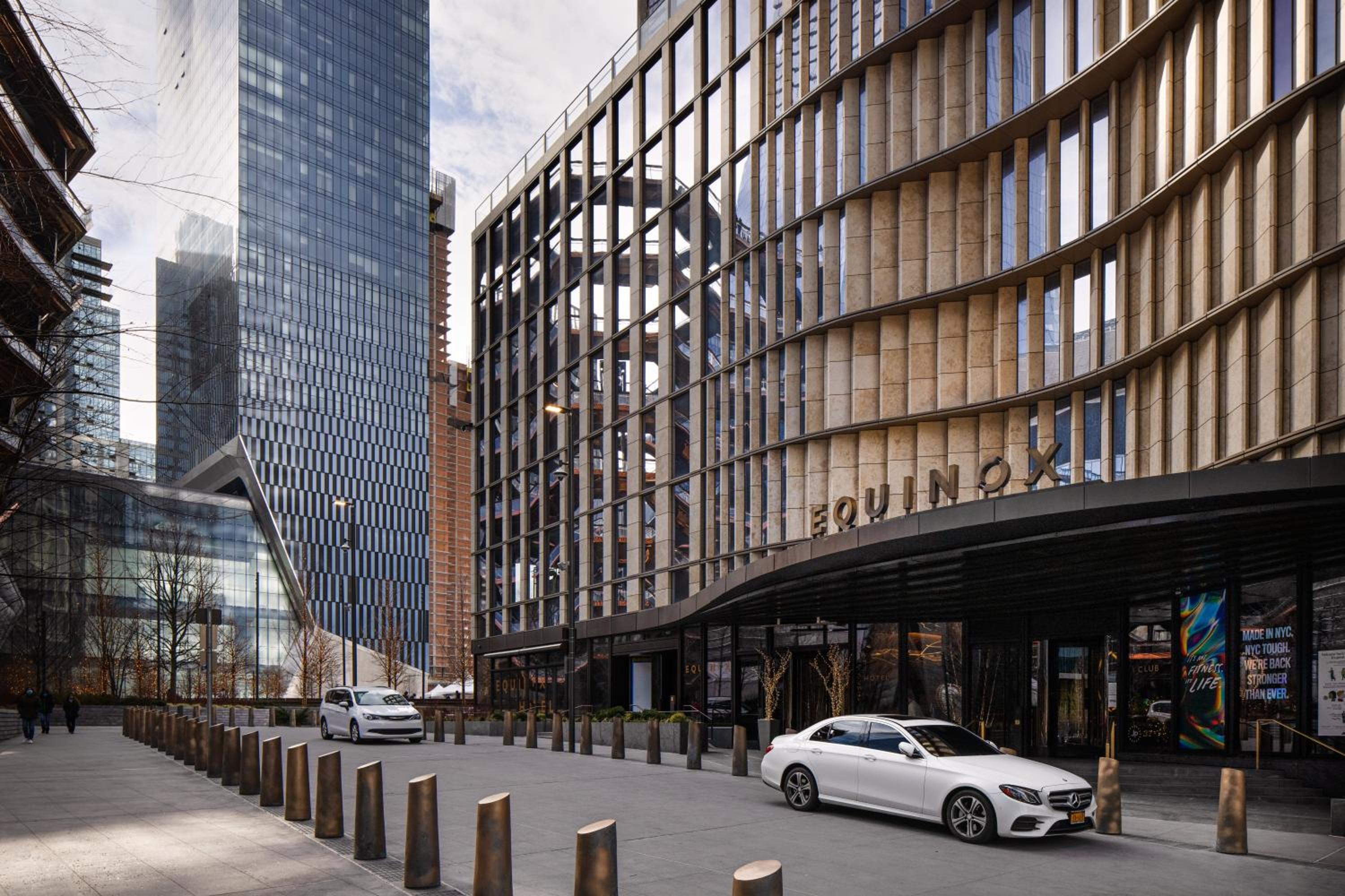 Sleek bronze lattework and geometric facade create striking street presence in Hudson Yards, where white sedans glide beneath the hotel's distinctive modernist canopy.