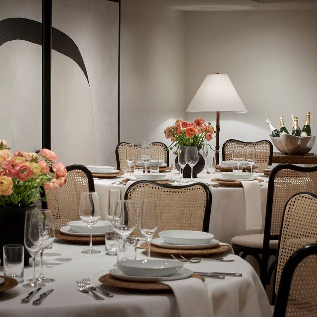 Cane-backed chairs and coral-hued ranunculus create intimate elegance in the dining room, where champagne chills nearby and soft lamplight warms cream-colored walls.