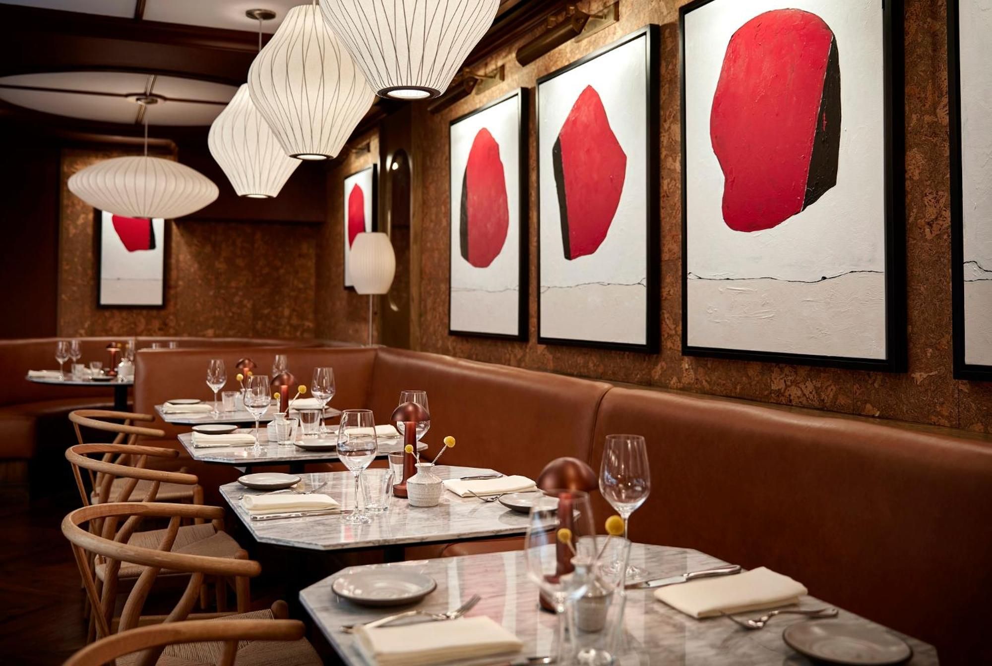 Warm wood beams frame bold red abstract art above banquette seating, where modern pendant lights cast an intimate glow over marble-topped tables set for dinner.