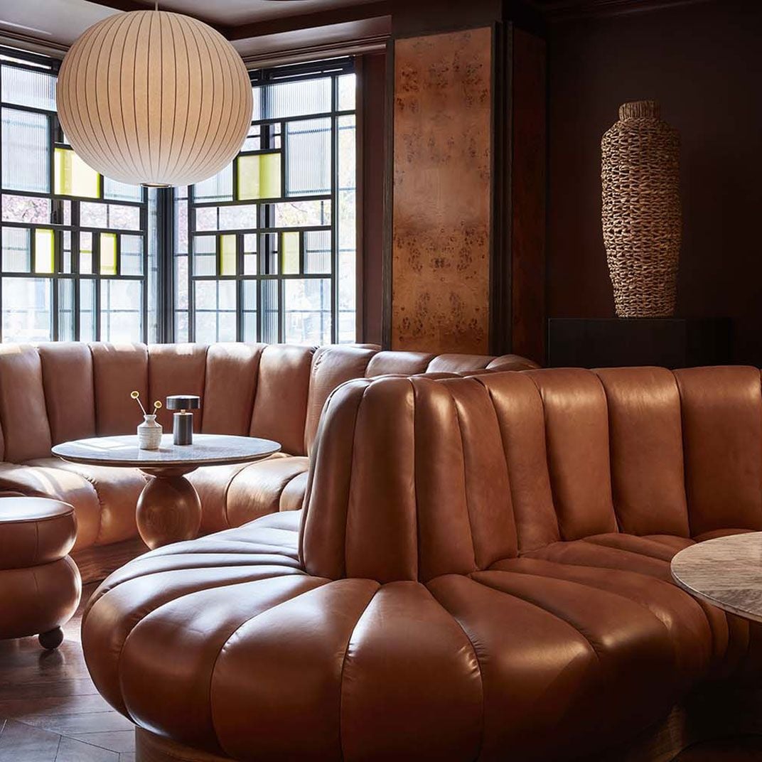 Sculptural cognac leather banquettes curve through the moody lounge, where ribbed pendant lights cast warm glows against rich wood paneling and textured wall art.