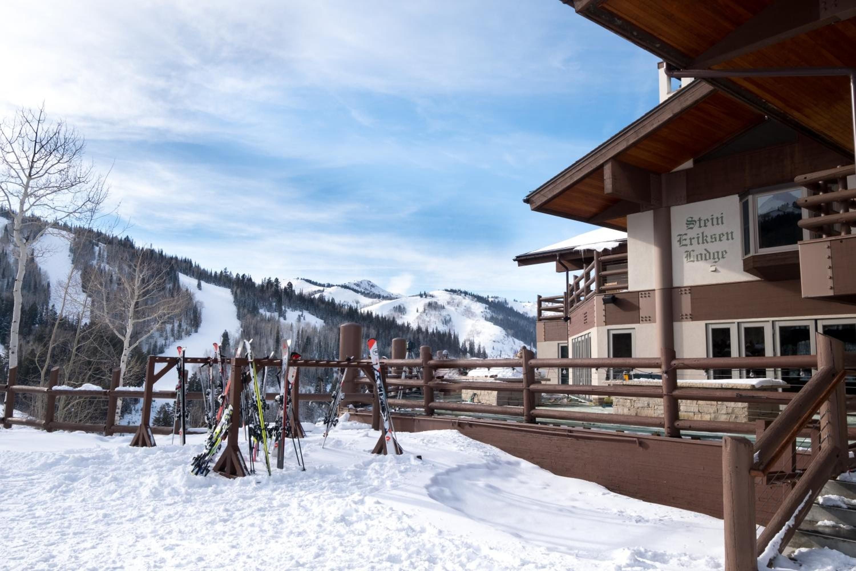 Alpine tranquility meets mountain luxury as ski equipment rests against wooden racks, framing powder-dusted slopes and the lodge's signature timber architecture beneath Utah's crystalline winter sky.