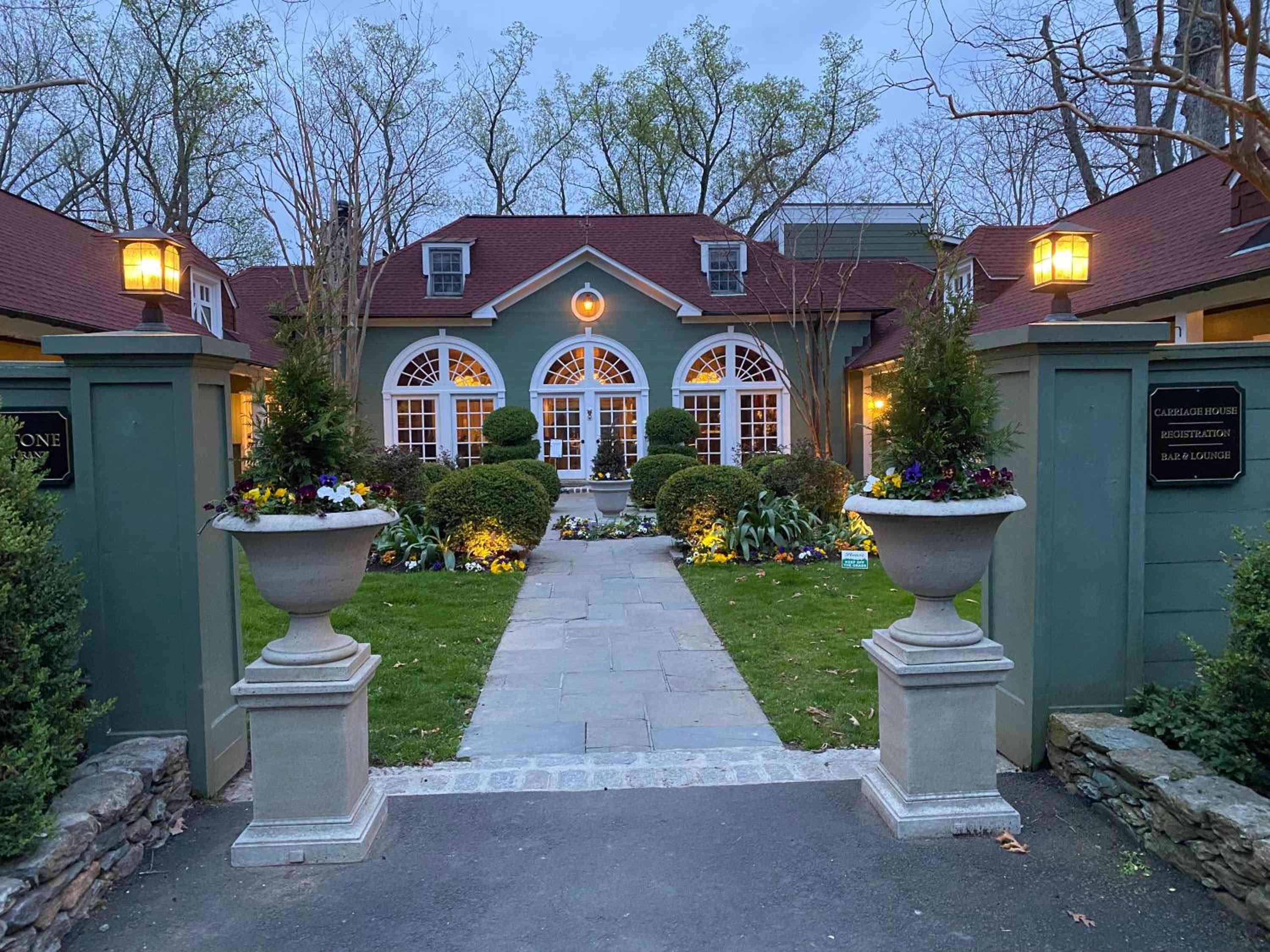 Stone urns brimming with seasonal blooms frame the torch-lit entrance, where arched windows glow warmly against sage-green siding and burgundy rooflines at dusk.
