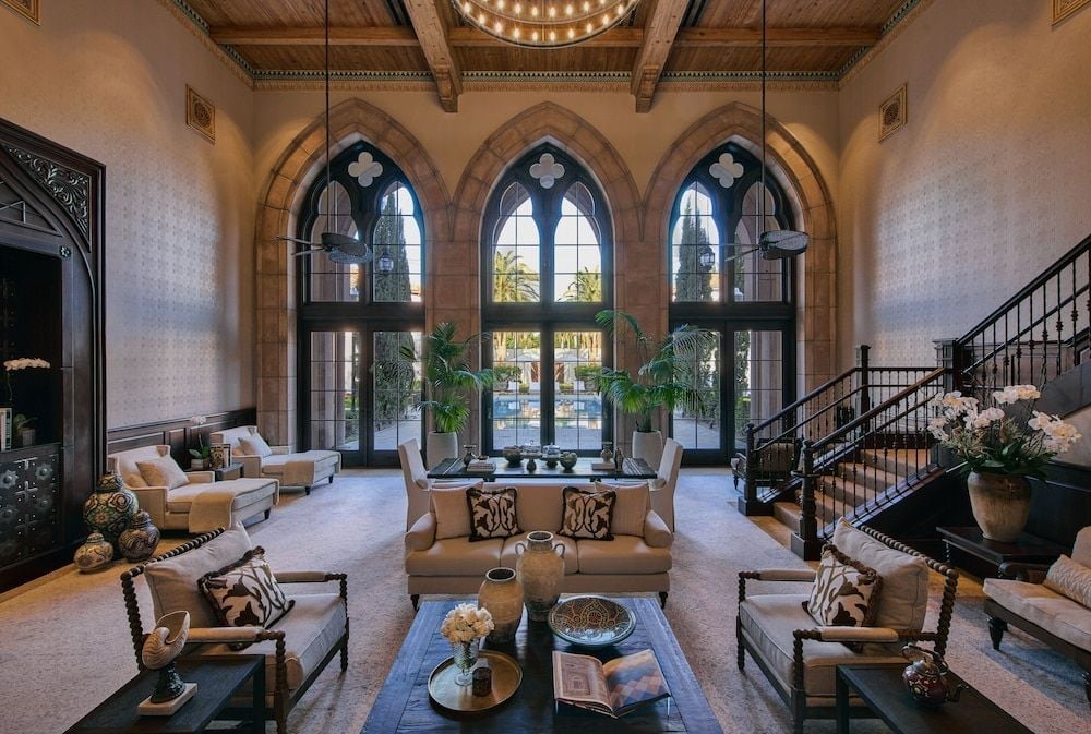 Gothic arches frame this dramatic double-height lobby where neutral furnishings and potted palms create intimate conversation areas beneath exposed wooden beams and wrought iron chandeliers.