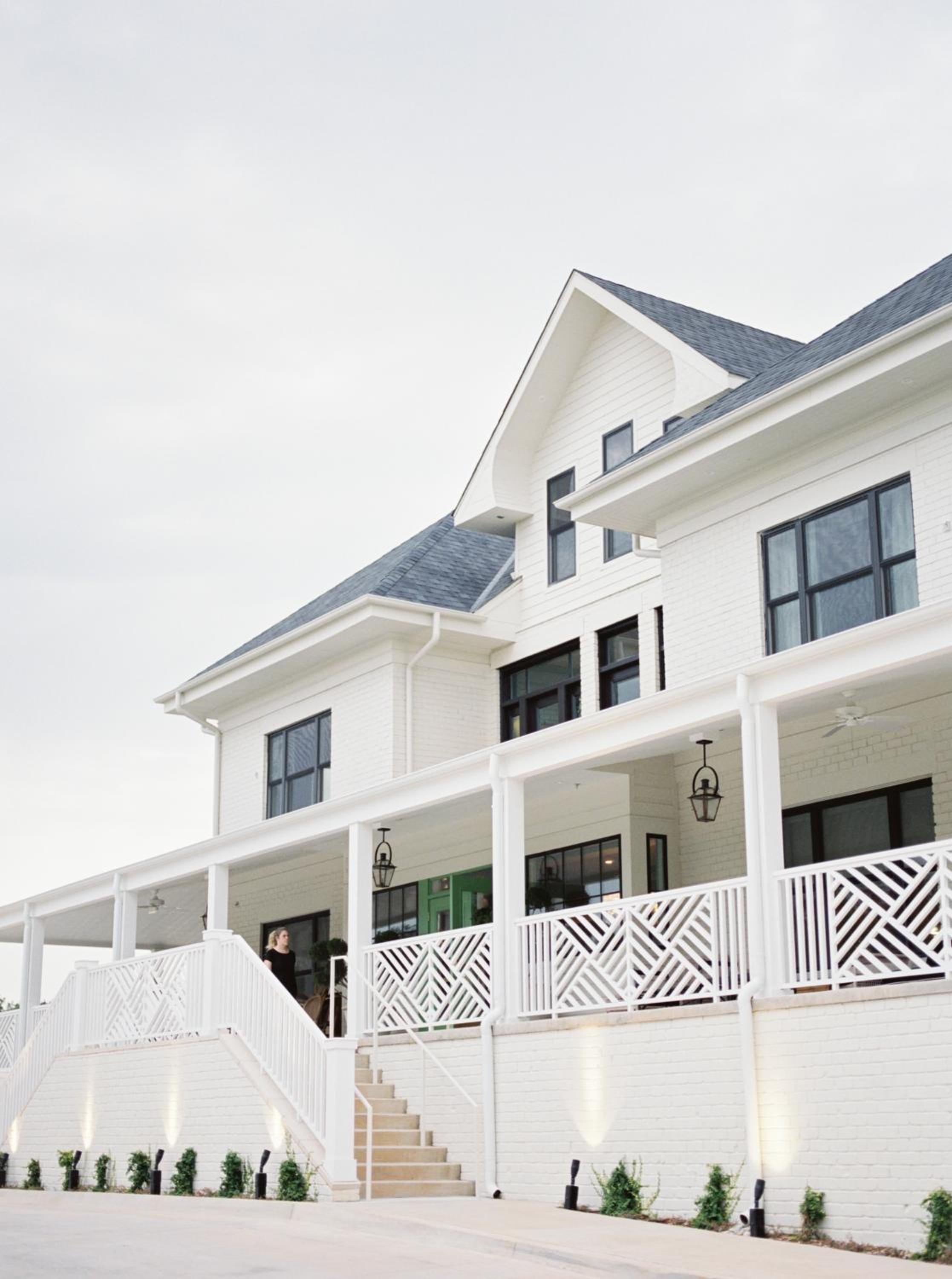 White clapboard siding and geometric porch railings create striking coastal elegance, while lantern fixtures cast warm light across the welcoming wraparound veranda.