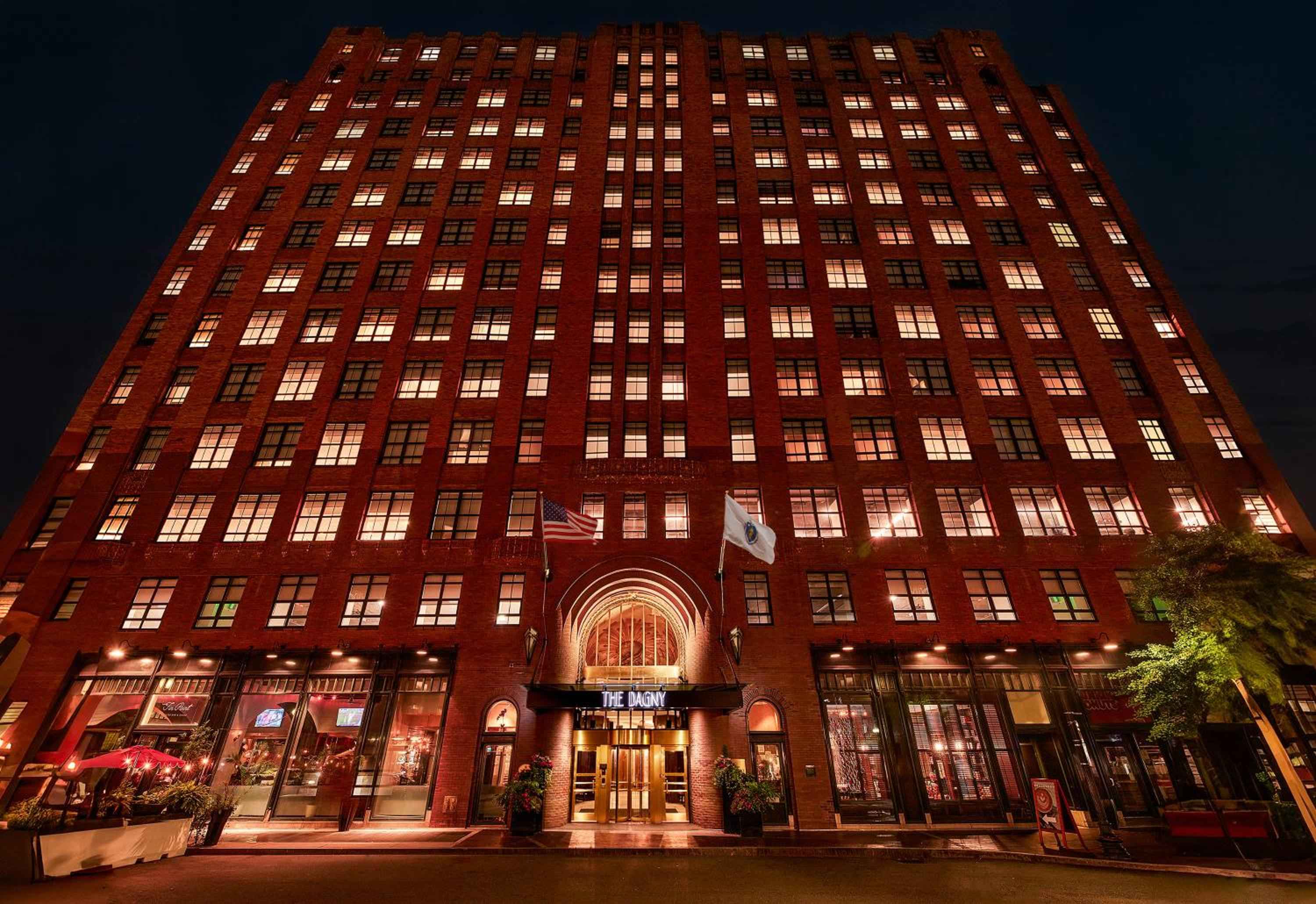 Warm golden light spills from countless windows across the red brick facade, while the illuminated arched entrance beckons beneath fluttering flags on this atmospheric Boston evening.