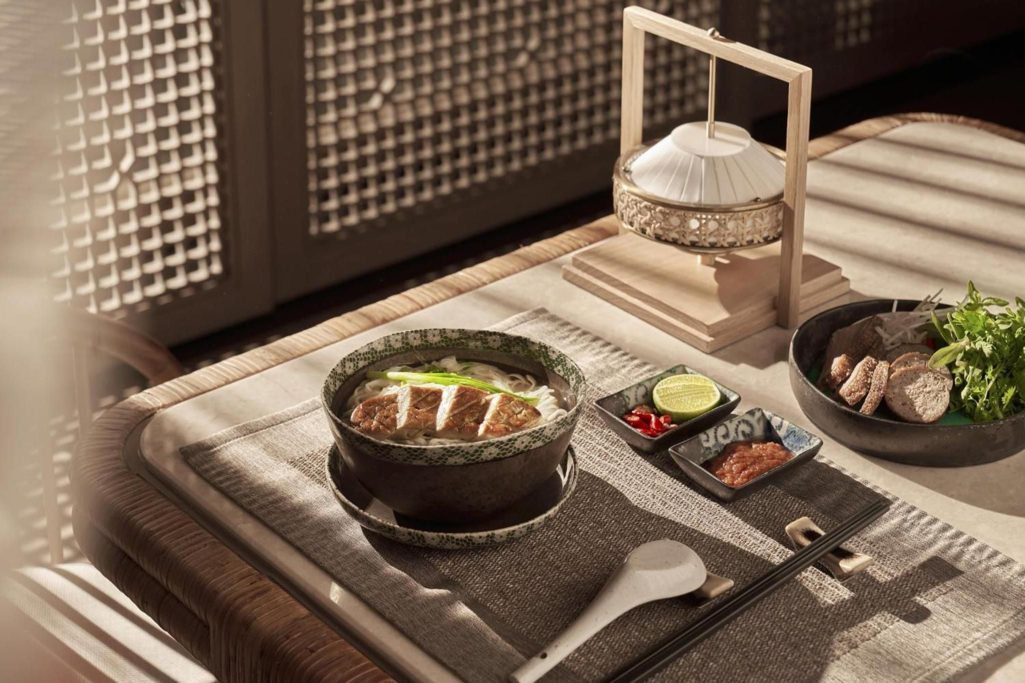 Traditional Vietnamese pho arrives steaming hot in handcrafted ceramics, accompanied by fresh herbs and house-made condiments served on woven bamboo placemats.