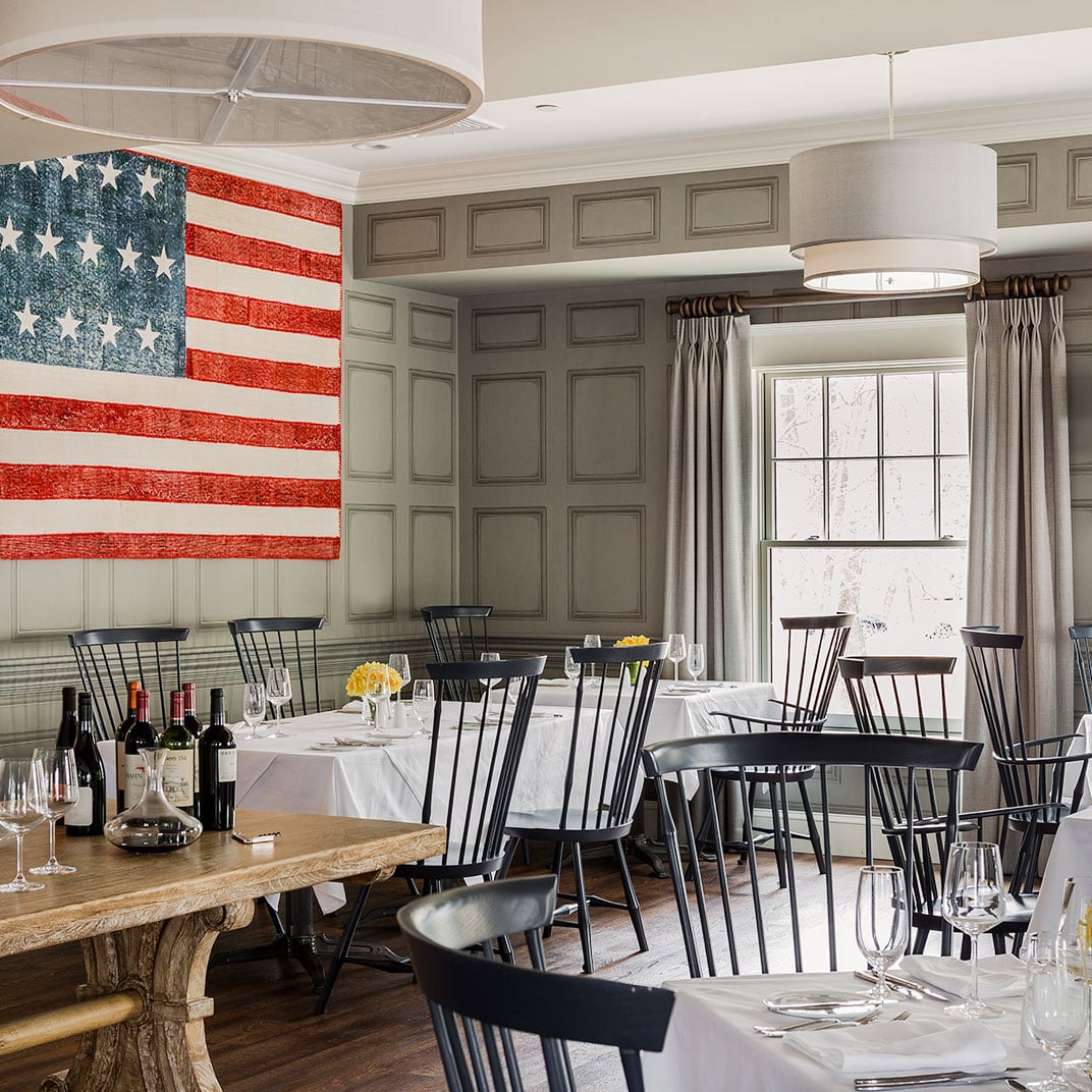 Windsor chairs and weathered farm tables create an intimate dining atmosphere beneath a vintage American flag, where colonial charm meets contemporary culinary excellence.