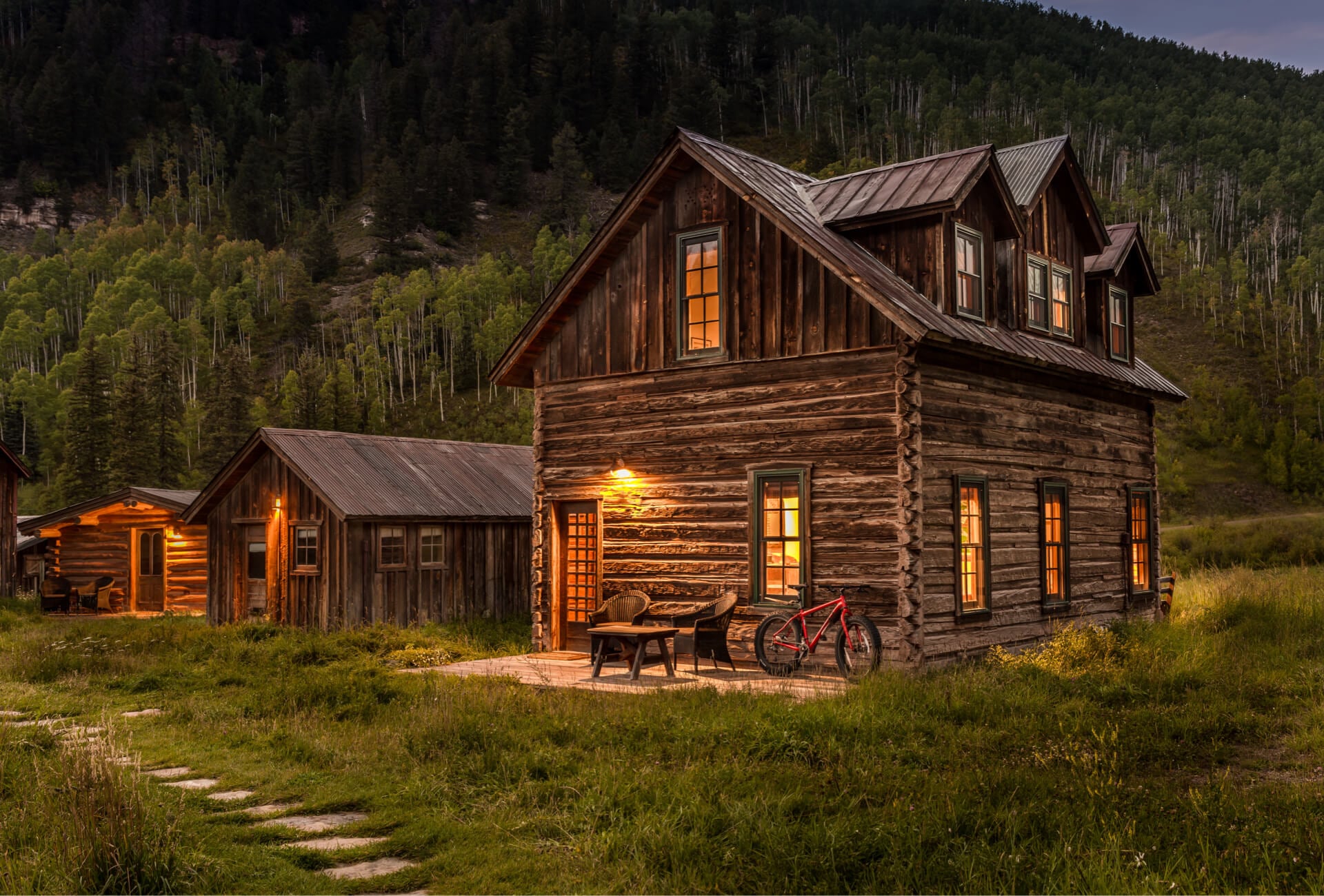 Weathered log cabins glow with golden lamplight against aspen-covered peaks, where vintage bicycles and worn wood tell stories of Colorado's mining past.